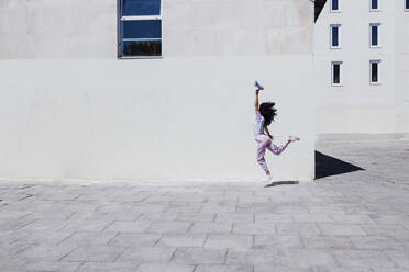 Junge Frau mit erhobener Hand, die ein Megaphon hält, während sie an einem sonnigen Tag durch ein Gebäude springt - EGHF00012