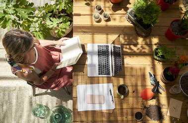 Mature woman reading diary while sitting with laptop at table in backyard - VEGF04670