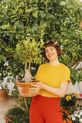 Smiling mid adult woman holding Ficus Microcarpa plant in home garden - MGRF00283