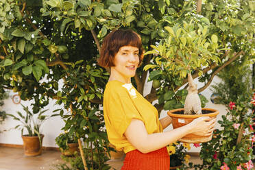 Smiling woman holding Ficus Microcarpa plant by tree in backyard - MGRF00284