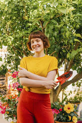 Smiling woman with hand tool standing in home garden - MGRF00285