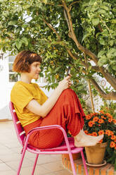 Frau benutzt ihr Smartphone, während sie auf einem Stuhl im heimischen Garten sitzt - MGRF00287