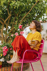 Smiling woman with eyes closed holding tea mug on chair in home garden - MGRF00290