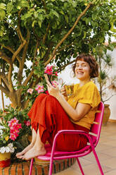 Lächelnde Frau mit Mobiltelefon beim Tee trinken auf einem Stuhl im heimischen Garten - MGRF00292