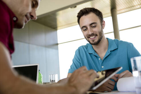 Mittlerer erwachsener Geschäftsmann, der über ein digitales Tablet mit einem lächelnden männlichen Kollegen im Büro diskutiert - BMOF00841