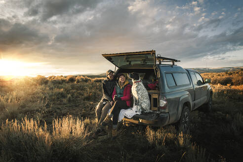 Cheerful couple sitting with dog at trunk of off-road vehicle on field during vacation - CAVF94213