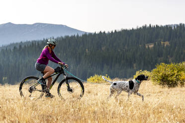 Seitenansicht einer lächelnden Frau, die im Urlaub mit dem Fahrrad hinter einem Hund auf einem Feld fährt - CAVF94225