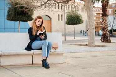 Young woman writes in her notebook seated on a stone bench - CAVF94278