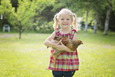 Cute little blond girl with blue eyes holding a chicken outdoors. - CAVF94295