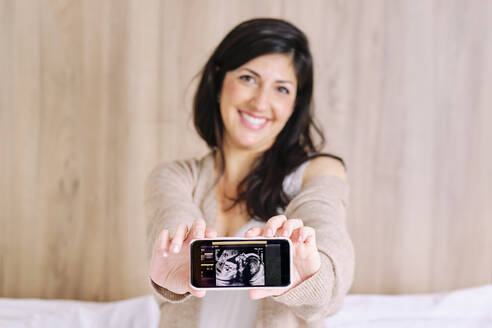 Smiling pregnant woman showing ultrasound on mobile phone - AODF00508