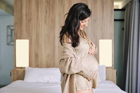 Smiling pregnant woman with hands on stomach standing in bedroom at home - AODF00513