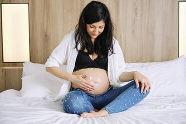 Pregnant woman applying moisturizer on belly while sitting on bed in bedroom - AODF00522