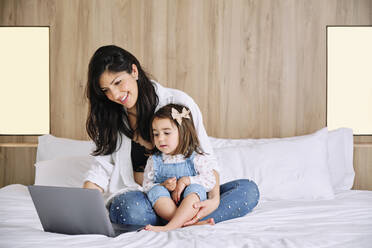 Smiling mother using laptop while sitting with daughter on bed at home - AODF00525