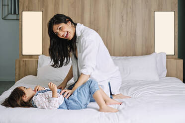 Cheerful mother and daughter playing over bed at home - AODF00526