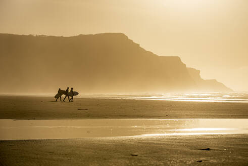 Menschen mit Surfbrettern gehen am Strand entlang im Abendsonnenlicht, Rhossili, Gower Peninsula, Swansea, Wales, Vereinigtes Königreich, Europa - RHPLF19817