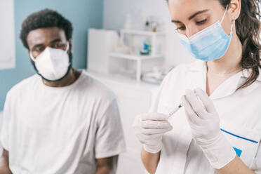 Ärztin mit Latexhandschuhen und Gesichtsschutz, die eine Spritze aus einer Impfstoffflasche einfüllt und sich darauf vorbereitet, einen nicht erkennbaren afroamerikanischen Mann zu impfen, der während des Ausbruchs des Coronavirus in einer Klinik behandelt wird - ADSF25162