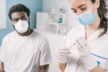 Female doctor in latex gloves and face shield filling in syringe from bottle with vaccine preparing to vaccinate unrecognizable African American man patient in clinic during coronavirus outbreak - ADSF25162