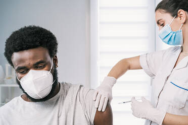 Female medical specialist in protective uniform, latex gloves and face mask vaccinating African American man patient in clinic during coronavirus outbreak - ADSF25163