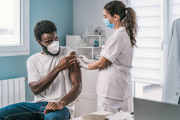 Medizinische Fachkraft in Schutzkleidung, Latexhandschuhen und Gesichtsmaske bei der Impfung eines afroamerikanischen Mannes in einer Klinik während des Ausbruchs des Coronavirus - ADSF25165