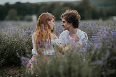 Gelassenes Paar, das in einem Lavendelfeld mit blühenden Blumen sitzt und sich gegenseitig anschaut - ADSF25187