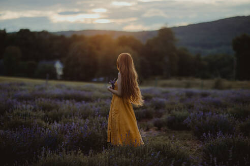 Seitenansicht einer zarten Frau mit Blumen in der Hand, die in einem blühenden Lavendelfeld steht und die Natur genießt, während sie wegschaut - ADSF25188