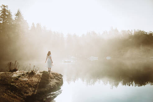 Rückenansicht Frau in einem weißen Kleid auf einem Felsen stehend mit Blick auf einen See an einem nebligen Tag - ADSF25189