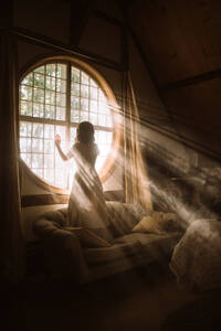 Rückenansicht einer nicht erkennbaren Frau in einem Kleid, die auf einer Couch vor einem runden Fenster in einem Haus an einem sonnigen Tag steht - ADSF25192