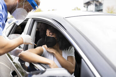 Side view of male doctor in protective uniform, latex gloves and face shield vaccinating African American female patient inside the car on a drive through mobile clinic during coronavirus outbreak - ADSF25206