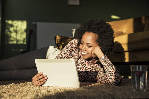 Lächelnde Frau, die ein digitales Tablet benutzt, während sie auf einem Teppich liegt - UUF23531