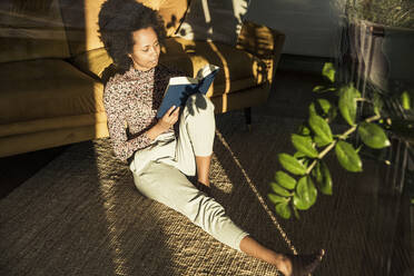 Woman reading book while sitting on carpet at home - UUF23533