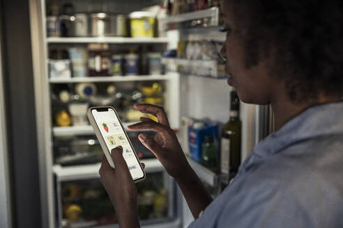 Frau, die zu Hause am Kühlschrank per Smartphone die Kalorien von Lebensmitteln zählt - UUF23565