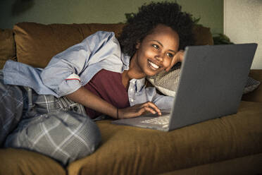Junge Frau benutzt einen Laptop, während sie zu Hause auf dem Sofa liegt - UUF23571