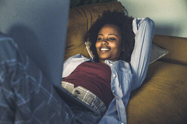 Lächelnde Frau, die einen Laptop benutzt, während sie sich auf dem Sofa zu Hause ausruht - UUF23573