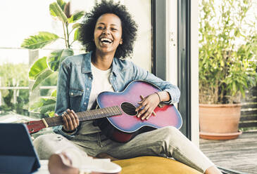 Fröhliche Frau spielt Gitarre, während sie zu Hause sitzt - UUF23580