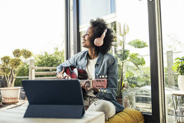 Woman listening music through headphones while playing guitar at home - UUF23584