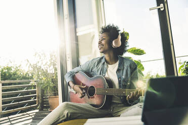 Lächelnde Frau hört Musik über Kopfhörer, während sie zu Hause Gitarre spielt - UUF23585