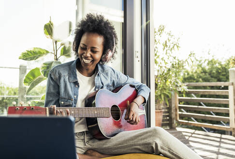 Fröhliche Frau mit Kopfhörern spielt Gitarre, während sie zu Hause mit einem digitalen Tablet lernt - UUF23586