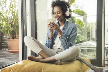 Smiling woman listening music through headphones while using mobile phone at home - UUF23588