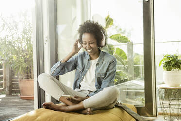 Cheerful woman wearing headphones using mobile phone at home - UUF23590