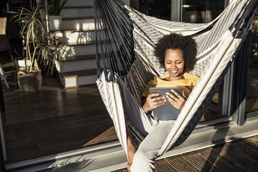 Lächelnde Frau, die ein digitales Tablet benutzt, während sie sich in der Hängematte an einem sonnigen Tag entspannt - UUF23596