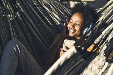 Smiling woman with headphones and mobile phone lying on hammock - UUF23599