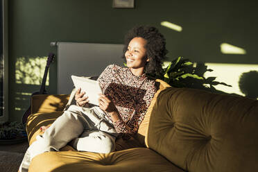 Junge Frau mit digitalem Tablet, die wegschaut, während sie auf dem Sofa sitzt - UUF23619