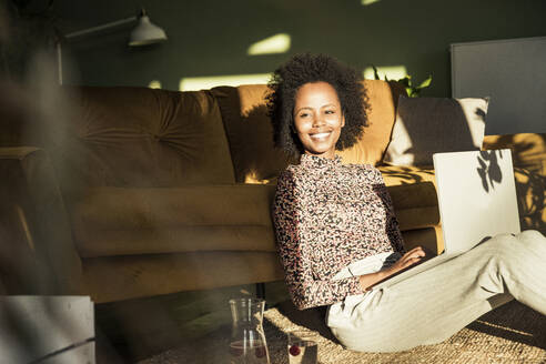 Junge Frau lächelt, während sie mit einem Laptop im Wohnzimmer sitzt - UUF23625