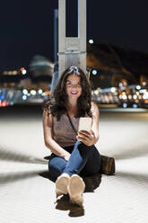 Smiling woman with mobile phone sitting on floor at pier during night - DLTSF01924