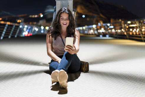 Lächelnde Frau, die ein Mobiltelefon benutzt, während sie nachts auf dem Pier sitzt - DLTSF01926