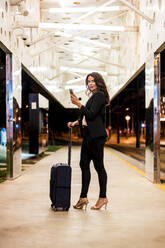 Weibliche Fachkraft mit Gepäck schaut weg, während sie an einer nächtlichen Straßenbahnhaltestelle steht - DLTSF01929