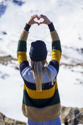 Junge Frau macht Herzform vor schneebedecktem Berg - JSMF02401