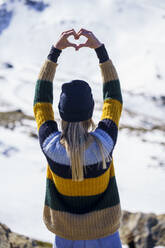 Young woman making heart shape in front of snowcapped mountain - JSMF02401