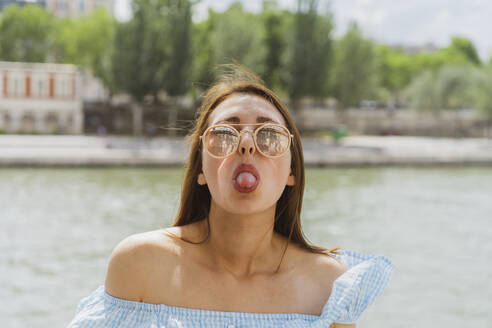 Frau mit Sonnenbrille streckt vor einem Fluss die Zunge heraus - AFVF08957