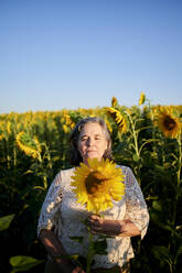 Ältere Frau mit geschlossenen Augen, die eine Sonnenblume auf einem Feld hält - KIJF03998
